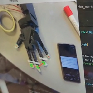 A robotic hand lies on a table with colored markers on its fingertips, while a smartphone and red marker pen sit nearby. A screen overlay shows finger positions being tracked in real-time using gesture recognition software, with lines and labels indicating finger joints and movements. A portion of code is visible on the side